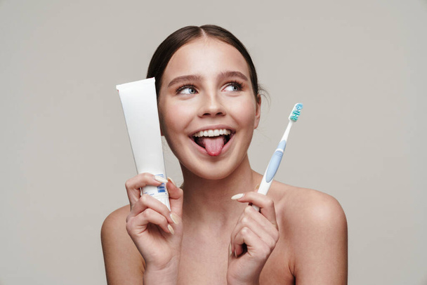 Image of funny shirtless woman smiling while holding toothpaste and toothbrush isolated over grey background - Foto, afbeelding