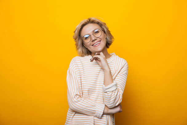 Monochrome photo of caucasian blonde woman wearing eyeglasses and smiling at camera touching her chin on a yellow studio wall - Photo, Image