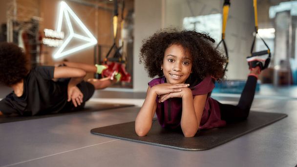 Teenage girl smiling at camera while doing exercises on mat using fitness straps in gym. Sport, healthy lifestyle, physical education concept - Foto, Imagen