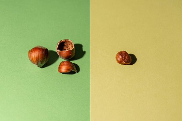 Oloupaný lískový ořech spolu se skořápkami a další lískový ořech ve skořápce na zeleném a žlutém pozadí - Fotografie, Obrázek