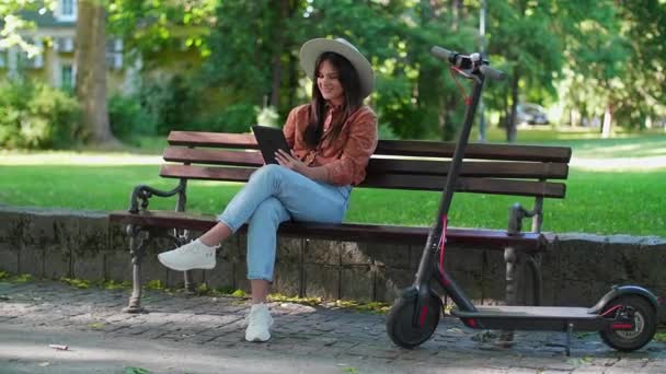 Una joven con un sombrero se sienta en un banco del parque y se desplaza en la tableta en sus manos. Un scooter eléctrico está aparcado junto a ella, mientras que los árboles y los colores verdes predominan en el fondo. - Imágenes, Vídeo