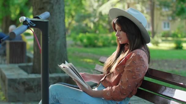 帽子を被った若い美少女がコーヒーを飲みながら本を手に公園のベンチに座っている。電動スクーターは彼女の隣に駐車されていますが、木はバックグラウンドで優勢です。. - 映像、動画