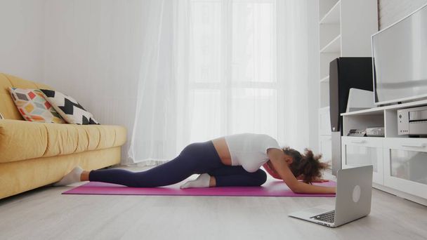 On-line pracy w domu kobieta korzystająca z usług internetowych z pomocą instruktora na laptopie w domu. Szczupła, sportowa Afroamerykanka ćwicząca jogę na macie. Młoda kobieta ćwiczy jogę w pokoju - Zdjęcie, obraz