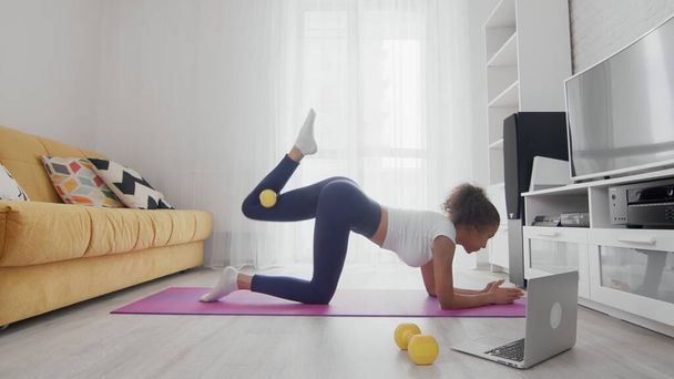 On-line home work out γυναίκα που χρησιμοποιεί υπηρεσίες διαδικτύου με τη βοήθεια του εκπαιδευτή της στο laptop στο σπίτι. Slim σπορ αφρικάνικη αμερικανική γυναίκα άρση αριστερό πόδι με κίτρινο αλτήρα στο χαλί - Φωτογραφία, εικόνα