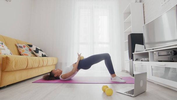 On-line home work out woman using internet services with help of her instructor on laptop at home. Slim sporty african american woman lifting pelvis with yellow dumbbell on mat - Photo, Image