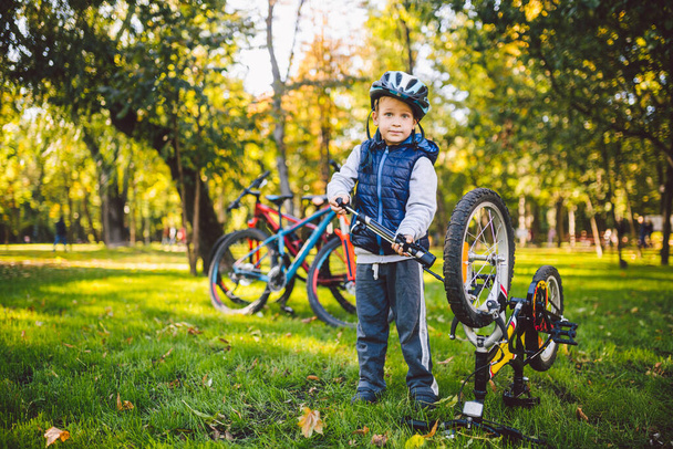 Enfant caucasien garçon dans un casque apprend à réparer son vélo. Enfant cycliste vérifie le mécanisme d'un vélo dans une clairière d'herbe verte dans le parc. Thème club sportif pour enfants. Section VTT
. - Photo, image