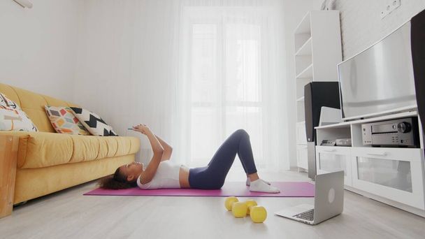 On-line home work out woman using internet services with help of her instructor on laptop at home. Slim sporty african american woman lays down on mat and uses phone after exercises at home - Photo, Image