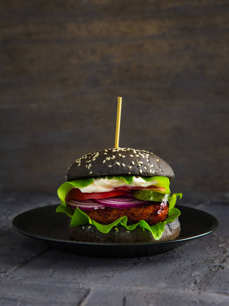 Schwarzer Burger mit Tofu, Salat, Tomate, Zwiebel und Gurke auf schwarzem Hintergrund.  - Foto, Bild