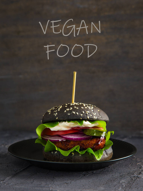 Hamburguesa negra con tofu, ensalada, tomate, cebolla y pepino sobre fondo negro, inscripción comida vegana
. - Foto, Imagen