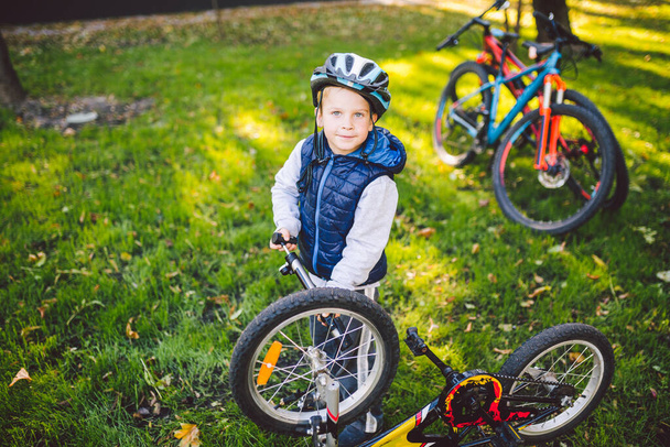 Белый мальчик в шлеме учится ремонтировать свой велосипед. Детский велосипедист проверяет механизм велосипеда на поляне зеленой травы в парке. Спортивный клуб тема для детей. Горная секция
. - Фото, изображение