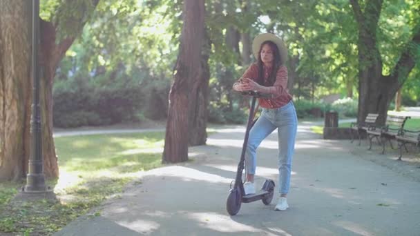 Una giovane bella donna con un cappello in piedi accanto al suo scooter elettrico mentre digita sul suo telefono. Sullo sfondo predominano alberi e colori verdi. - Filmati, video