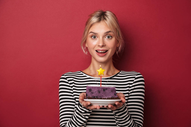 Image of nice delighted woman in striped sweatshirt holding cake with candle isolated over red background - Foto, imagen