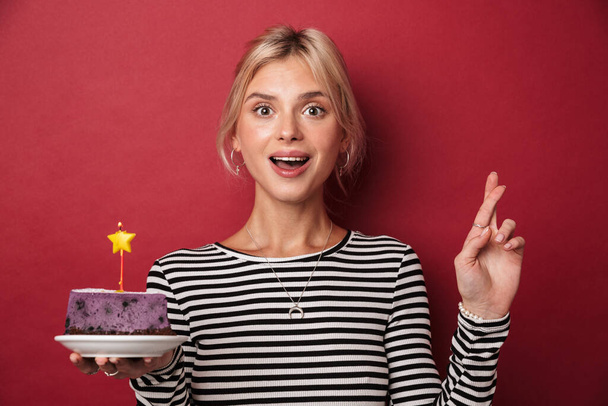 Imagen de una agradable mujer excitada sosteniendo el pastel y sosteniendo los dedos cruzados para una buena suerte aislada sobre fondo rojo
 - Foto, imagen