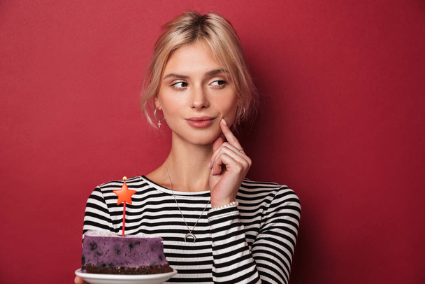 Image of nice thinking woman in striped sweatshirt holding cake with candle isolated over red background - Foto, imagen