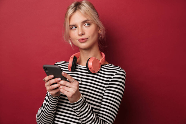 Image of nice thinking woman with headphones typing on cellphone isolated over red background - Foto, Bild