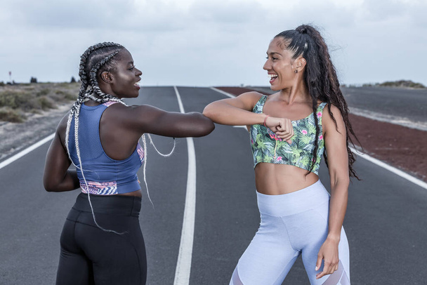 Позитивні різноманітні жінки в спортивному одязі посміхаються і дивляться один на одного, стоячи на дорозі і підстрибуючи лікті під час сеансу на околиці
 - Фото, зображення