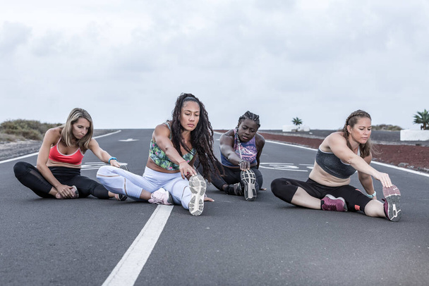 Atletas femeninas multiétnicas de cuerpo completo en ropa deportiva inclinándose hacia adelante y tocando los dedos de los pies mientras están sentadas en la carretera y calentándose durante el entrenamiento en las afueras
 - Foto, imagen