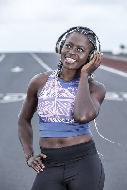 Positive African American female smiling and looking up while listening to favorite music in headphones during break in outdoor workout - Photo, Image