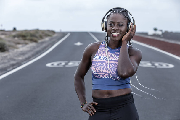 Deportista negra positiva con la mano en la cintura sonriendo y escuchando música en auriculares mientras descansa en la carretera durante la sesión de running en las afueras
 - Foto, Imagen