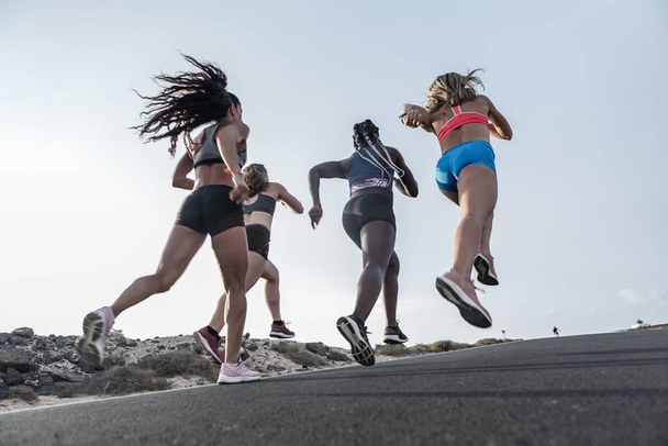 Névtelen, többnemzetiségű női sportruhás barátok rohangálnak aszfaltozott úton a felhők nélküli esti égbolton a külvárosi futás közben - Fotó, kép