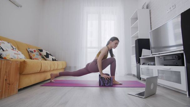Он-лайн тренировать женщину используя интернет-услуги с помощью своего инструктора. Женщина делает упражнения на фиолетовом коврике и смотрит на блокнот
 - Фото, изображение