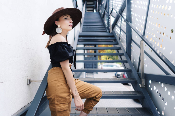 Модна брюнетка з довгим волоссям, одягнена в стильний великий плетений капелюх, позує на сходах
 - Фото, зображення
