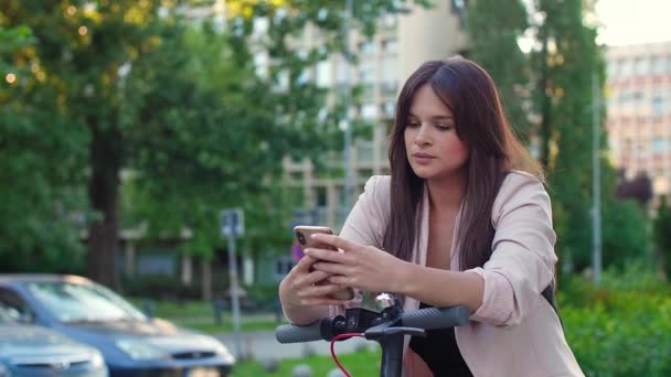 Egy fiatal, gyönyörű nő áll az elektromos robogó mellett, miközben gépel a telefonján. Komoly, üzletszerűen öltözött, modern építészettel a háttérben.. - Felvétel, videó