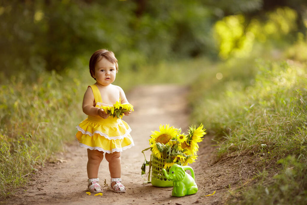 Roztomilá holčička se slunečnicí. Dítě s květinou. Jeden rok starý. Stojí v lese na stezce. Nedaleko je zelená zavlažovací plechovka, kytice slunečnic. Vypadá to jako zlatíčko. Žluté šaty. - Fotografie, Obrázek