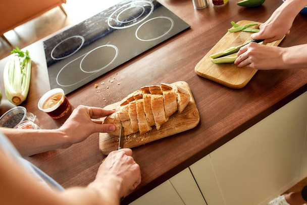 Cropped shot of couple cutting tasty homemade bread and slicing cucumber while preparing healthy meal in the kitchen together. Vegetarianism, healthy food concept - Photo, Image