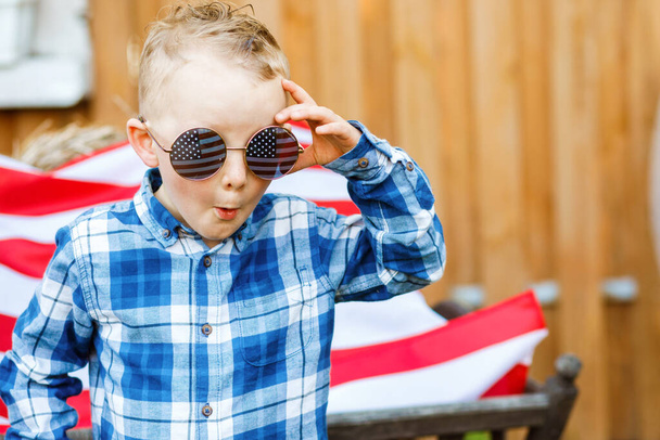 スタイリッシュなガラスのかわいいブロンドの男の子は、アメリカの国旗の背景に庭に立っています - 写真・画像