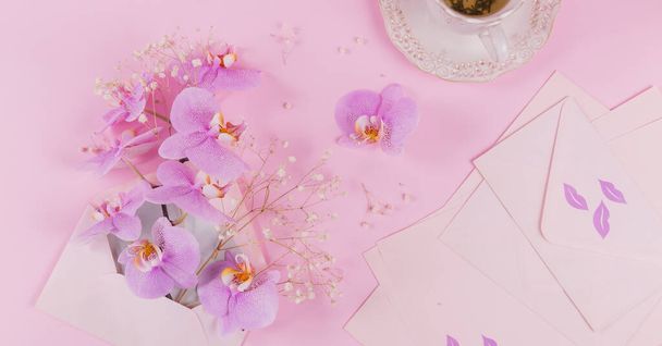Delicate top view composition with morning cup of tea, pink letter bag full of purple orchid flowers and empty envelop on light pink background. Beautiful breakfast surprise concept - Фото, изображение
