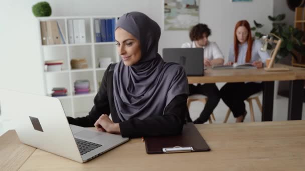 Muslim woman drinking coffee and having a video talk on a laptop in the office - Materiaali, video