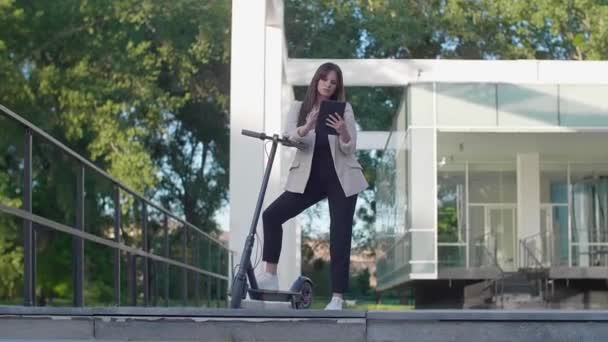 Una joven y hermosa mujer está de pie junto a su scooter eléctrico, con un pie en una plataforma de pie. Ella sonríe, entrenada en negocios, con la arquitectura moderna en el fondo. - Imágenes, Vídeo