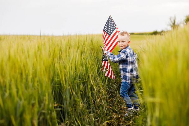 Un chico rubio lindo está parado en un campo con una bandera americana en sus manos
. - Foto, Imagen