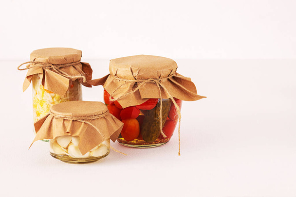 gefermenteerde groenten in transparante glazen potten bedekt met papier van gerecycleerde materialen. tomaten, knoflook, kool. Op een witte roze achtergrond. kopieerspaaten - Foto, afbeelding