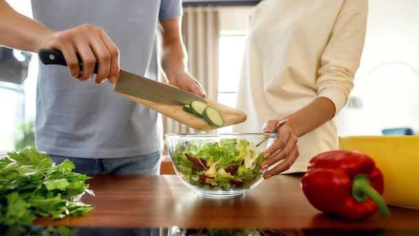 Gehakt schot van een man die gesneden komkommer in een schotel deed terwijl een vrouw hem hielp, met de salade schotel in haar hand. Vegetariërs bereiden samen gezonde maaltijd in de keuken - Foto, afbeelding