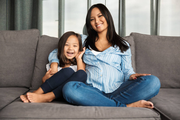 Photo of happy pregnant asian woman with her little daughter hugging and laughing while sitting on couch at home - Zdjęcie, obraz