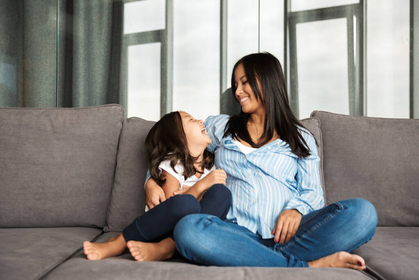 Photo of happy pregnant asian woman with her little daughter hugging and laughing while sitting on couch at home - Fotó, kép