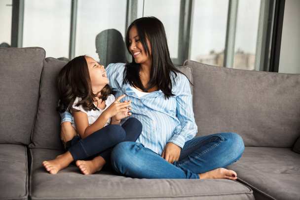 Photo of happy pregnant asian woman with her little daughter hugging and laughing while sitting on couch at home - Фото, зображення