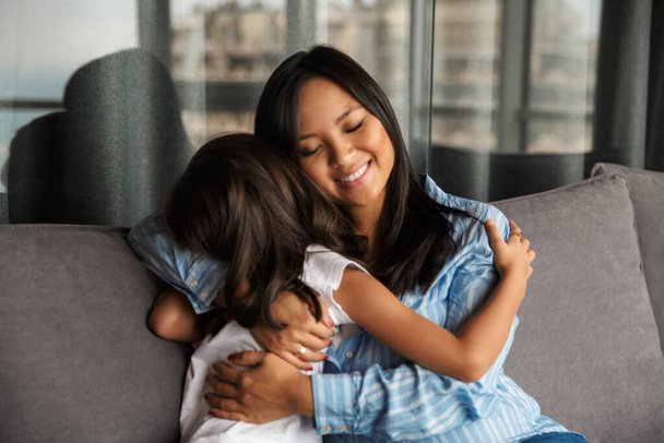 Photo of pleased pregnant asian woman with her little daughter hugging and smiling while sitting on couch at home - Φωτογραφία, εικόνα