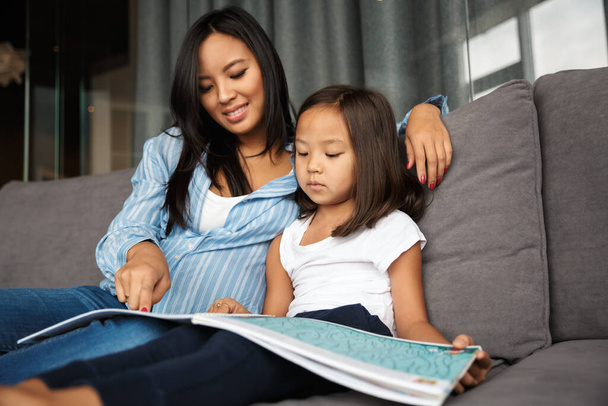 Photo of pleased pregnant asian woman with her little daughter reading book while sitting on couch at home - Photo, Image