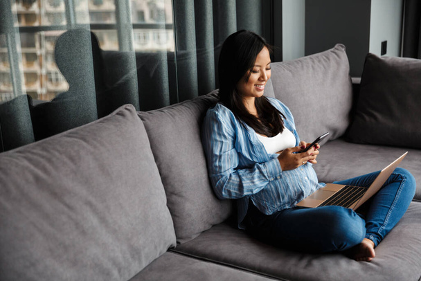 Photo of smiling pregnant asian woman using laptop and mobile phone while sitting on couch at home - Fotografie, Obrázek
