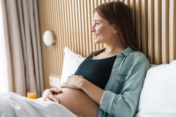 Photo of happy pregnant woman with red hair smiling and petting her belly while lying on bed in sunny room - Фото, зображення