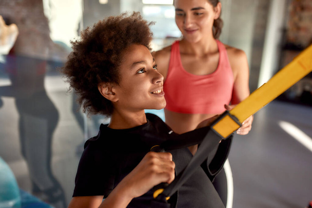 Portrait of teenage boy training using fitness straps in gym while female trainer helping him. Sport, healthy lifestyle, physical education concept - Foto, afbeelding