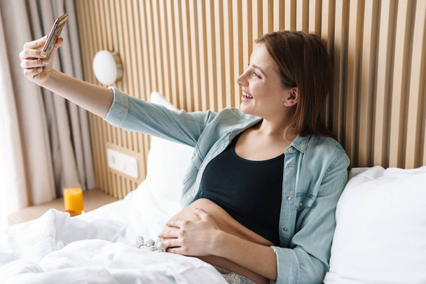 Photo of laughing pregnant woman with red hair taking selfie photo on mobile phone while lying on bed in sunny room - Photo, image