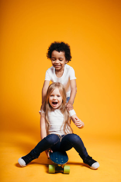 Curly dark-skinned boy rides his little girlfriend on a longboard, and the girl laughs on an orange background - Fotoğraf, Görsel