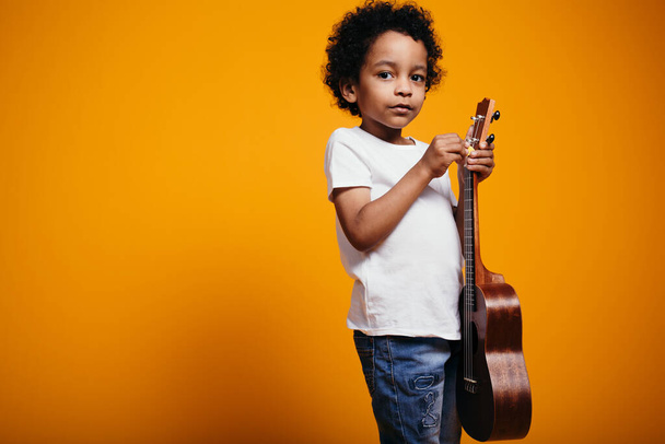 A little black boy holds a ukulele guitar in his hands and pulls up the strings so that they sound right - Zdjęcie, obraz