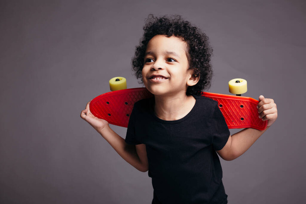 Little black boy in a black T-shirt holds a red longboard behind his back and smiles in the studio - Valokuva, kuva