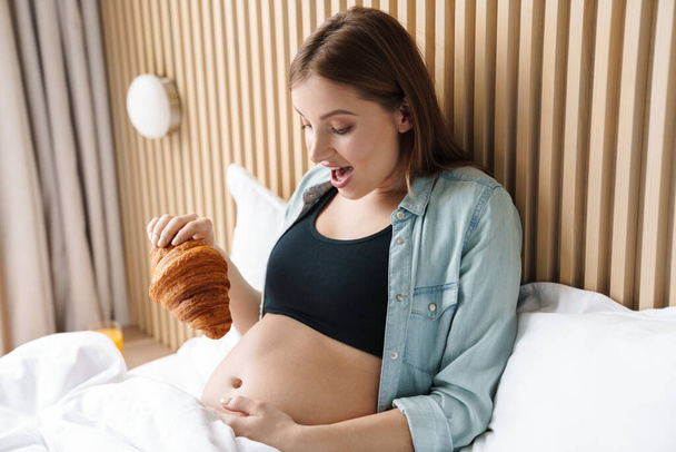 Photo of excited pregnant woman making fun with croissant while having breakfast in bed at sunny room - 写真・画像