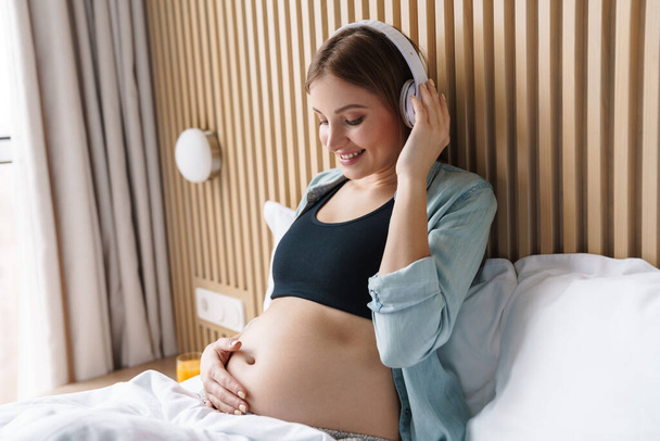 Photo of happy pregnant woman smiling and using wireless headphones while sitting in bed at sunny room - Foto, Bild